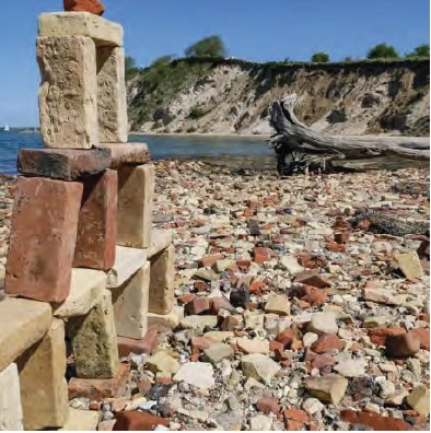 A small cairn of weathered ballast bricks stacked on a pebble beach, with a chalk cliff and driftwood in the background.