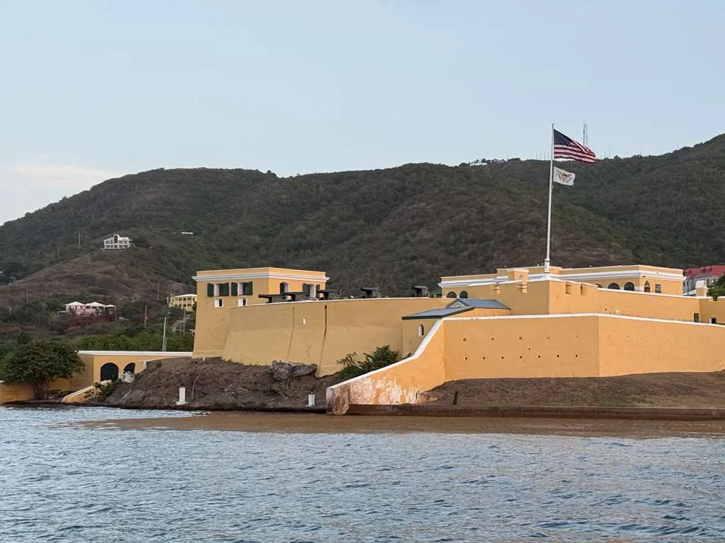 Fort Christiansvaern in Christiansted, St. Croix: a yellow Danish colonial fortification on the waterfront, flying the United States and Virgin Islands flags, with green hills rising behind.