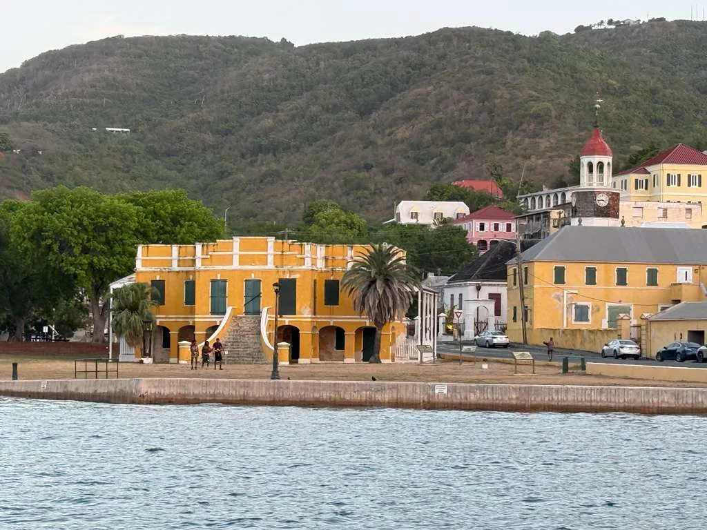 The yellow Danish West India and Guinea Company warehouse on the Christiansted waterfront, St. Croix, with the historic town, red-roofed buildings and the clock tower rising on the hillside behind it.