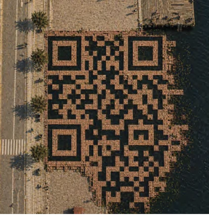 Straight-down aerial view of the memorial. The brick grid resolves into a complete QR code, with the three square finder patterns clearly visible at its corners, bounded by the promenade on one side and dark harbor water on the other.
